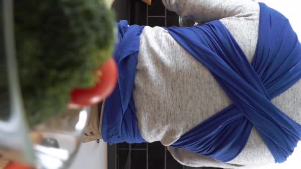垂直录像 爸爸带着小宝宝睡在厨房的吊带里做饭 男子休育儿假 做家务活 爸爸有照顾孩子的责任 — 图库视频影像