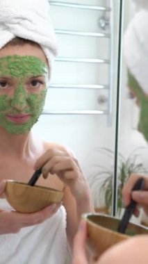 Yüzüne beyaz havlu takmış genç bir kadın yeşil nemlendirici bir maske takmış kameraya gülümsüyor ve komik bir yüz ifadesi takınıyor.