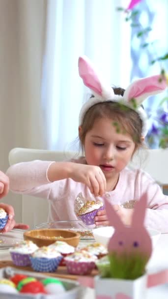 可爱的小女孩戴着滑稽的兔子耳朵 年轻的妈妈则坐在餐桌旁做复活节纸杯蛋糕 桌上放着篮子 鸡蛋和兔子 — 图库视频影像