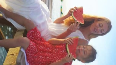 Mutlu anne ve kızı ahşap bir iskele üzerinde oturan ve gün ışığında sulu kırmızı karpuz yeme.