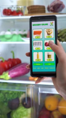 Çok yönlü video görüntüsü var. Akıllı Telefon Uygulaması Kullanarak İnternetten Yemek Siparişi Vermek. Ön taraftaki açık buzdolabında, cep telefonundaki kadın el işaretleme listesinin yakın çekimi. Gıda Teslimat Mobil Uygulaması.