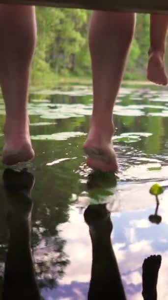 フィンランドの水面の近くに足を振る木の小屋の端に座っている森の湖の沈黙を楽しんでいる子供の若いカップル 垂直方向のビデオ — ストック動画