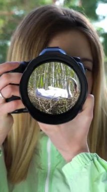 Göl kıyısında su kuşlarını fotoğraflayan bir kadın. Kamera lensinde beyaz kuğu yansıması. Akıllı telefon ekranı ve mobil aygıtlar için dikey video