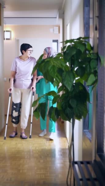 女性の介護者は 足の怪我で女性の患者を助け 手を握り 彼女を支えています ユニフォームでヒジャブを着用している若いムスリム看護師は 彼女が一緒に歩いている間 成熟した高齢女性をサポートしています — ストック動画