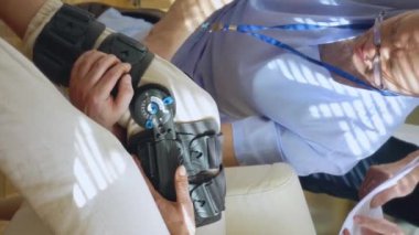 Erkek hemşire, klinikteki ya da hastanedeki uzman doktorla iletişim kuran yaşlı bir kadın hastanın bacağına kölelik, tıbbi destek ve dizlik takmaya yardım eder.