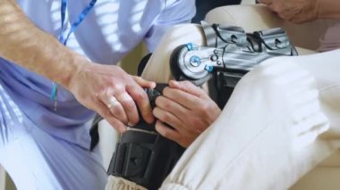 Erkek hemşire, klinikteki ya da hastanedeki uzman doktorla iletişim kuran yaşlı bir kadın hastanın bacağına kölelik, tıbbi destek ve dizlik takmaya yardım eder.