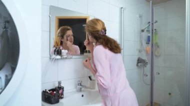 Güzel, beyaz, orta yaşlı bir kadın kaşlarını boyuyor, elinde fırçayla banyoda kaş modelliği yapıyor.