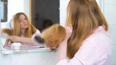 Çekici genç bir kadının banyoda aynanın yanında dikilirken güzel saçlarını tarayıp yansımasına baktığı arka plan. Saç bakımı kavramı.