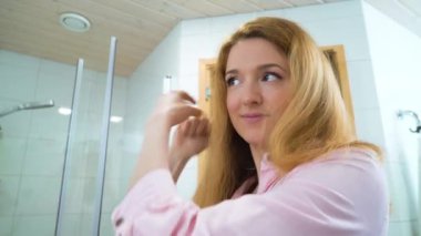 Çekici genç bir kadın banyoda aynanın yanında dikilirken güzel saçlarını tarayıp yansımasına bakıyor. Saç bakımı kavramı.