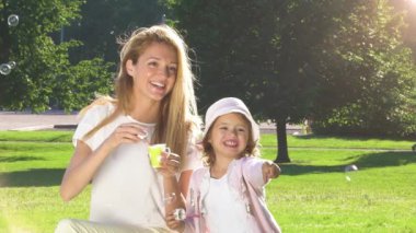 Genç anne ve tatlı kızı şehir parkında sabun köpüğüyle oynuyorlar. Yaz aile zamanı, baloncuk üfleme, güneşli bir hafta sonunda eğlenmek..