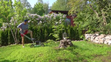 Orta yaşlı bir adam bahçesinde çim biçme makinesiyle fazla büyümüş yeşil çimenleri buduyor. Şortlu erkek bahçıvan bahçe düzenlerken çim biçme makinesi kullanıyor. Mevsimlik iş kavramı.