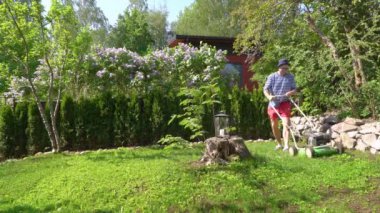 Orta yaşlı bir adam bahçesinde çim biçme makinesiyle fazla büyümüş yeşil çimenleri buduyor. Şortlu erkek bahçıvan bahçe düzenlerken çim biçme makinesi kullanıyor. Mevsimlik iş kavramı.