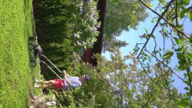 Orta yaşlı bir adam bahçesinde çim biçme makinesiyle fazla büyümüş yeşil çimenleri buduyor. Şortlu erkek bahçıvan bahçe düzenlerken çim biçme makinesi kullanıyor. Mevsimlik iş kavramı.