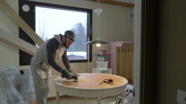 Koruyucu maskeli ve eldivenli orta yaşlı bir adam eski bir masanın yüzeyini öğütme makinesiyle öğütüyor. Mobilya yenileme, DIY onarım, kendi kendine tamir, geri dönüşüm, sürdürülebilir yaşam tarzı