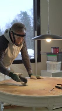 Koruyucu maskeli ve eldivenli orta yaşlı bir adam eski bir masanın yüzeyini öğütme makinesiyle öğütüyor. Mobilya yenileme, DIY onarım, kendi kendine tamir, geri dönüşüm, sürdürülebilir yaşam tarzı