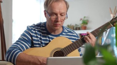 Orta yaşlı bir adam rahat bir odada oturuyor ve akustik gitar çalmayı öğrenmeye odaklanıyor. Önündeki küçük bir masaya yerleştirilmiş dizüstü bilgisayarında bir video dersi izliyor.