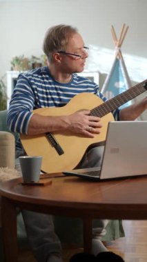 Orta yaşlı bir adam rahat bir odada oturuyor ve akustik gitar çalmayı öğrenmeye odaklanıyor. Önündeki küçük bir masaya yerleştirilmiş dizüstü bilgisayarında bir video dersi izliyor.