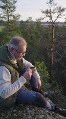 Orta yaşlı bir adam uçurumun tepesinde oturmuş kuzey ormanında günbatımının tadını çıkarıyor ve geleneksel Fin ahşap kuksa fincanından sıcak bir içecek içiyor.