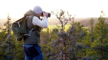 Orta yaşlı bir adam, el değmemiş bir Finlandiya ormanında uçurumun kenarında renkli bir günbatımına hayran olan bir kamerayla. Evergreen ağaçları, turuncu renkli gökyüzü, macera ve derin bir bağ.