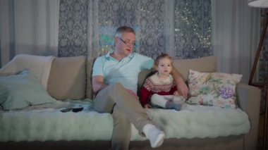 Orta yaşlı bir adam, yürümeye yeni başlamış çocuğuyla sıcak bir oturma odasında oturuyor ve akşamları televizyon seyrediyor. Oda, televizyonun parıltısıyla hafifçe aydınlatılıyor. Çocukların odaklanmış ifadesini vurguluyor..