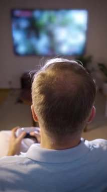 Akşamları konsolda video oyunu oynayan orta yaşlı bir adam modern oturma odasındaki rahat bir koltukta oturuyor ve arka planda televizyon ekranı parlıyor..