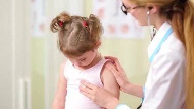 Beyaz laboratuar önlüklü bir kadın doktor grip mevsiminde steteskopla küçük bir kızın nefesini dinleyerek onu muayene ediyor..