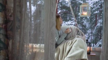 Sıcak kış yaşam tarzı. Mutlu genç kadın, verandada bir hamakta oturarak sıcak kahvenin tadını çıkarıyor. Kış ağacı arka planında balkon. Kişisel tecrit, ev karantinası..