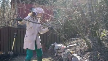 Açık hava iş kıyafeti, eldiven ve başörtüsü giymiş bir kadın bahar başında bahçedeki bir ağaç dalını temizliyor. Köy çitleri, ahşap bir ev ve doğal elementler.