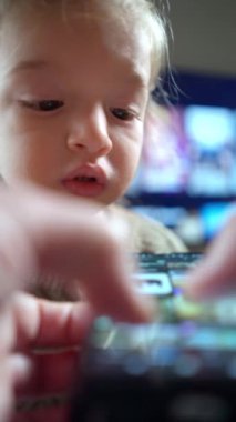 Çocuk akıllı telefon ekranı ile etkileşimde. Yetişkinlerin elleri ön planda cihazı kaydırıyor ve arka planda bir televizyon arayüzü var. Teknoloji, çocukluk, ekran süresi ve aile etkileşimi