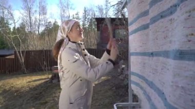 Açık renk ceketli ve çiçekli başörtülü bir kadın Finlandiya 'nın güneşli bahar bahçesinde klasik bir halıyı temizliyor veya dövüyor. Kırsal ya da banliyölerde bahar temizliği gelenekleri