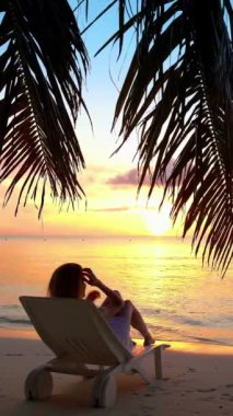 Günbatımında rahatlatıcı bir tropikal plaj sahnesi. Bir kadın kumlu sahildeki şezlongda uzanmış, palmiye yapraklarıyla çerçevelenmiş okyanusa bakıyor..