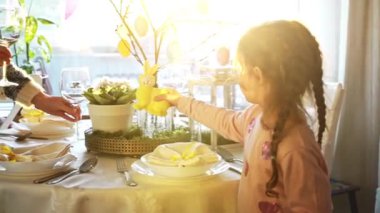 Küçük kız ve annesi, sarı tavşan desenli bayramlık Paskalya masası servis ediyorlar. Güneş ışığı konforlu odayı, zarif sofra takımlarını, pastel renkleri ve tatil dekorunu doldurur. Sevinç dolu sıcak bir aile anı