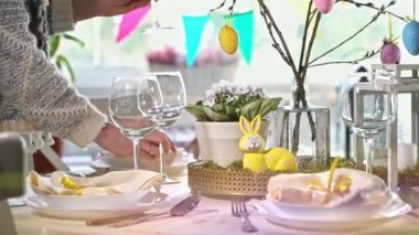 Genç bir kadın, sarı tavşan desenli bayramlık bir Paskalya masası servis ediyor. Güneş ışığı, zarif sofra takımları, pastel renkler ve bayram dekorunun altını çizerek, rahat odayı doldurur. Sıcak bir aile anı