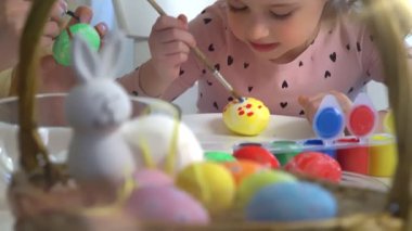 Anne ve kızı evde birlikte paskalya yumurtası boyuyor. Parlak şenlik dekorasyonları, neşeli atmosfer, yaratıcılık ve bağ. Renkli boyalar, tavşan kulakları ve bahar havası..