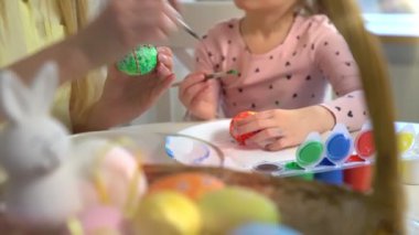 Anne ve kızı evde birlikte paskalya yumurtası boyuyor. Parlak şenlik dekorasyonları, neşeli atmosfer, yaratıcılık ve bağ. Renkli boyalar, tavşan kulakları ve bahar havası..