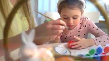 Anne ve kızı evde birlikte paskalya yumurtası boyuyor. Parlak şenlik dekorasyonları, neşeli atmosfer, yaratıcılık ve bağ. Renkli boyalar, tavşan kulakları ve bahar havası..