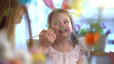 Annesi yüzünü boyarken tavşan kulaklı gülümseyen küçük bir kız. Sıcak, yaratıcı ve aile bağlarıyla dolu neşeli bir Paskalya anı. Yumuşak ışıklandırma, renkli dekorasyonlar, şenlikli ve