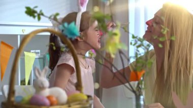 Annesi yüzünü boyarken tavşan kulaklı gülümseyen küçük bir kız. Sıcak, yaratıcı ve aile bağlarıyla dolu neşeli bir Paskalya anı. Yumuşak ışıklandırma, renkli dekorasyonlar, şenlikli ve