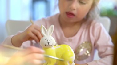 Anne ve kızı Paskalya için birlikte seramik bir tavşan boyuyorlar. Tavşan kulağı takan kız, annesi asistanlık yaparken dikkatlice sarı boya sürüyor. Masa renkli boyalarla dolu.