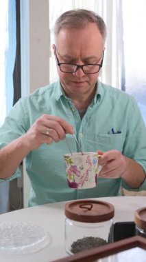 Bir adam çay içirerek organik çay demliyor. Beyaz masada, içinde çay olan cam kavanozlar ve tahta bir kutu var. Arkasında bitkiler ve raflar olan parlak, rahat bir mutfak..