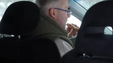Orta yaşlı, gözlüklü ve kışlık giysili bir adam ev yapımı bir sandviç yiyor ve karlı bir günde arabasında otururken tekrar kullanılabilir bir bardaktan kahve içiyor. Yolculuk sırasında hızlı bir yemekten hoşlanır.