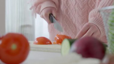 Genç kız parlak bir mutfakta taze bir salata hazırlıyor. Etrafı dolgu biber, salatalık ve pırasa gibi canlı malzemelerle çevrili bir tahtada sebze doğruyor. Konforlu, güneşli alan...