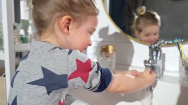 Genç bir çocuk banyo lavabosunda ellerini dikkatlice yıkıyor. Rahat pijamalar giyen çocuk, akan suyun altında sabunla iyi bir sabun yaratmaya odaklanmış..
