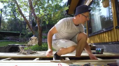 Güneşli bir yaz gününde bir adam sıcacık bir evin arka bahçesine ahşap bir teras inşa eder. Güverte üzerinde çalışırken bir matkap ve diğer aletleri kullanıyor. Yeşillik ve doğal ışıkla çevrili.