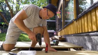 Güneşli bir yaz gününde bir adam sıcacık bir evin arka bahçesine ahşap bir teras inşa eder. Güverte üzerinde çalışırken bir matkap ve diğer aletleri kullanıyor. Yeşillik ve doğal ışıkla çevrili.