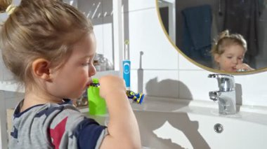 Banyoda küçük bir çocuk aynaya bakarken dişlerini fırçalar. Elinde bir diş fırçası ve yeşil bir kupa tutuyor. Parlak ve modern ev ortamında iyi hijyen alışkanlıkları ediniyor..