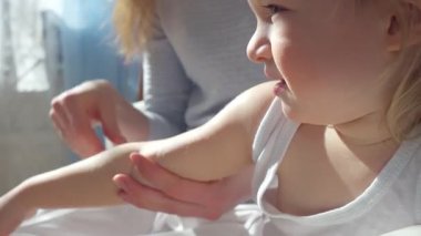Anne, çocuğun ellerine ve ayaklarına doğal ışık altında nemlendirici krem sürüyor. Nazik cilt bakımı, ebeveyn-çocuk bağı ve sağlıklı cilt koruması. Yakın çekim, sıcak atmosfer, kişisel bakım.