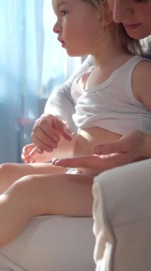 Anne, çocuğun ellerine ve ayaklarına doğal ışık altında nemlendirici krem sürüyor. Nazik cilt bakımı, ebeveyn-çocuk bağı ve sağlıklı cilt koruması. Yakın çekim, sıcak atmosfer, kişisel bakım.