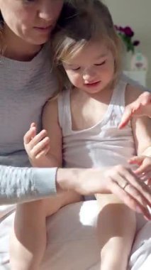 Anne, çocuğun ellerine ve ayaklarına doğal ışık altında nemlendirici krem sürüyor. Nazik cilt bakımı, ebeveyn-çocuk bağı ve sağlıklı cilt koruması. Yakın çekim, sıcak atmosfer, kişisel bakım.