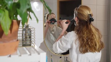 Beyaz bornozlu genç bir kadın parlak bir banyoda yüzünde siyah bir maske çıkarıyor. Doğal ışık ve modern ev dekorasyonu ile rahatlatıcı cilt bakımını seviyor..